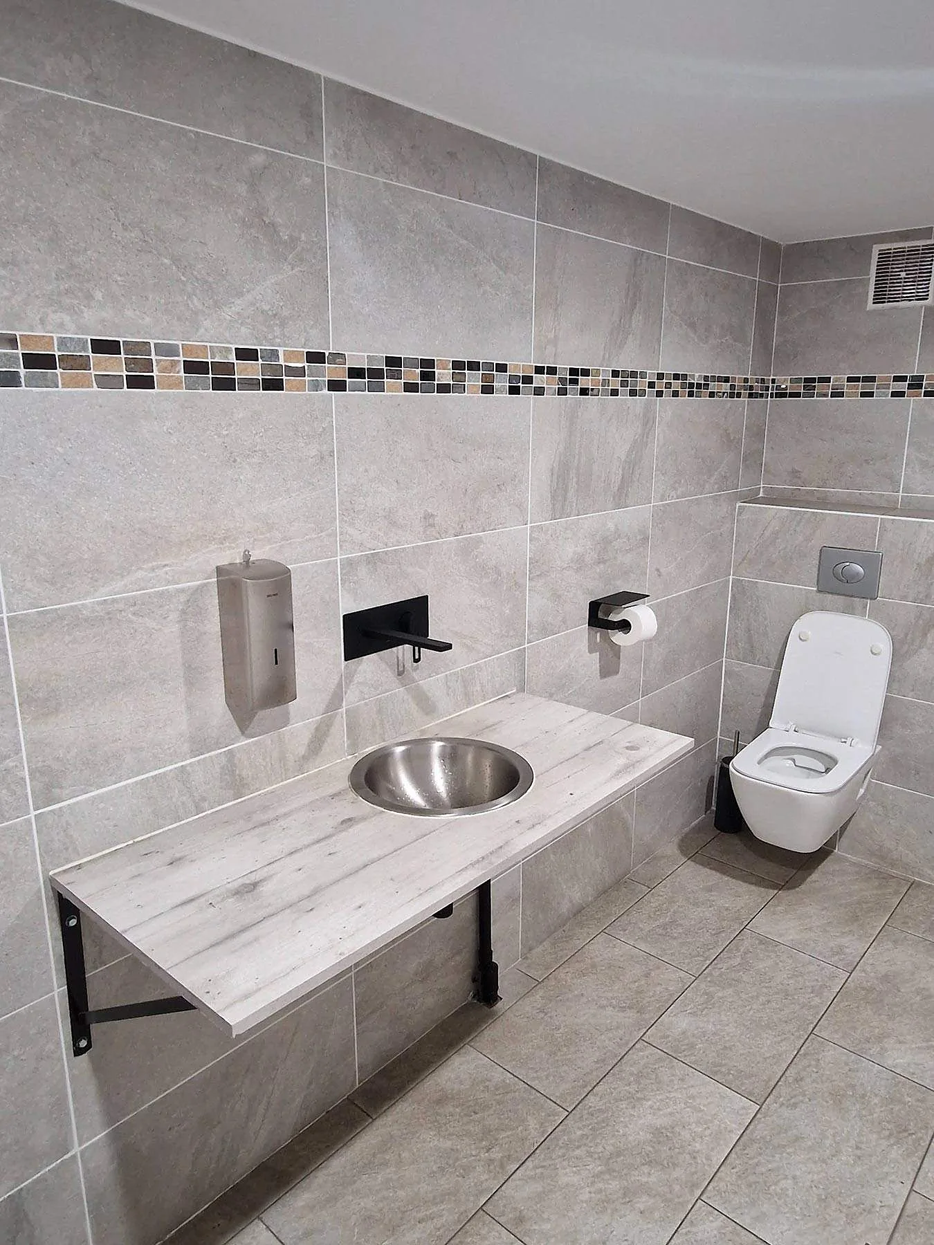 Une salle de bains équipée : plan vasque en inox, robinet mural noir et WC suspendu
